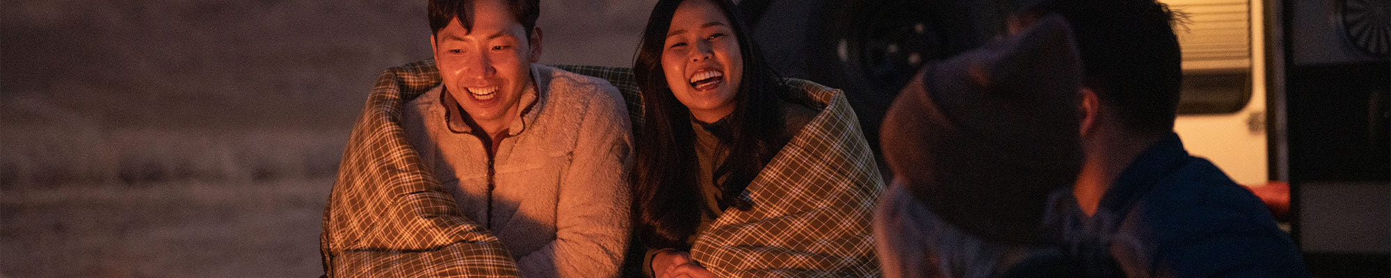 A couple laughs together by a campfire while snuggled up in a TETON Sports Evergreen Mammoth Sleeping Bag.