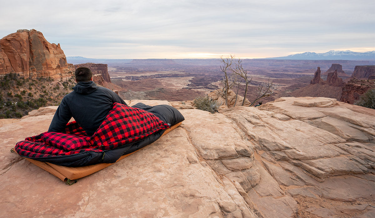 Aa man in a TETON Sports Celsius XXL Sleeping Bag sitting on a red rock cliff. His back is to the camera and he is gazing out on the desert scene below him.