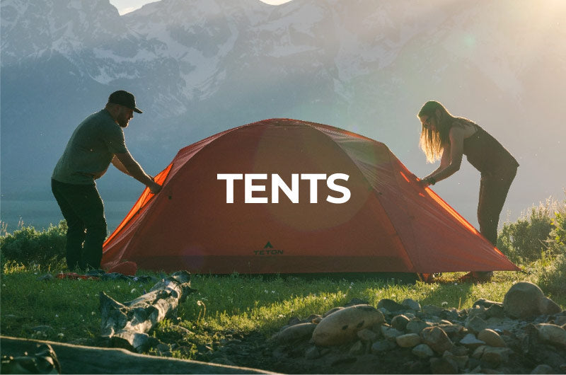 Shop Tents: Image shows a man and a woman working together to set-up a red TETON Sports Mountain Ultra tent in a camp site that has mountains in the background.